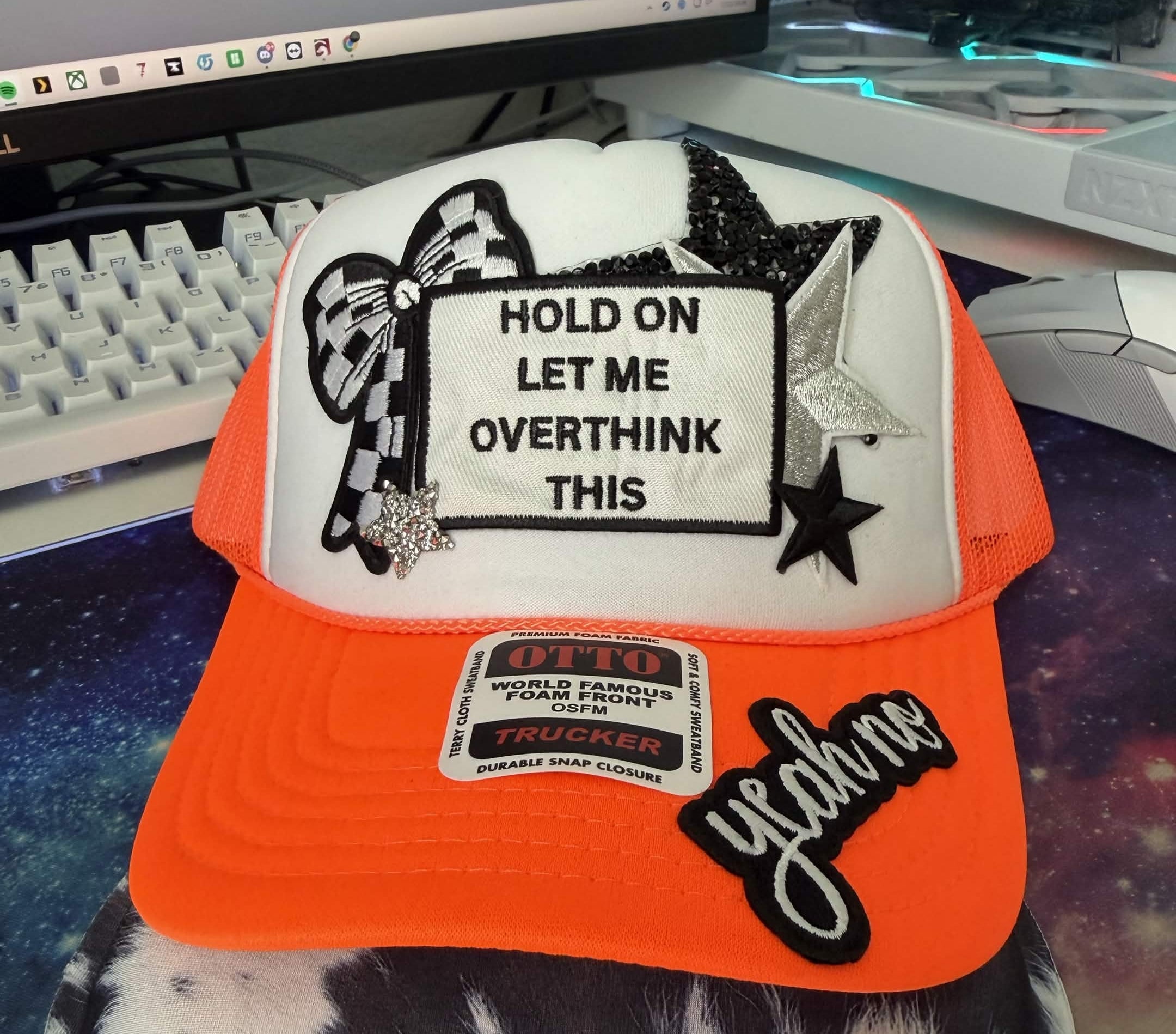 A custom trucker hat with an orange visor, white front panel, and black mesh sides. The front panel features a white patch with black text reading 'HOLD ON LET ME OVERTHINK THIS' surrounded by black and glittery star and bow graphics.