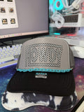 A gray and black trucker hat with a turquoise chain detail and a large checkered 'HOWDY' graphic on the front, displayed on a desk in front of a computer monitor.