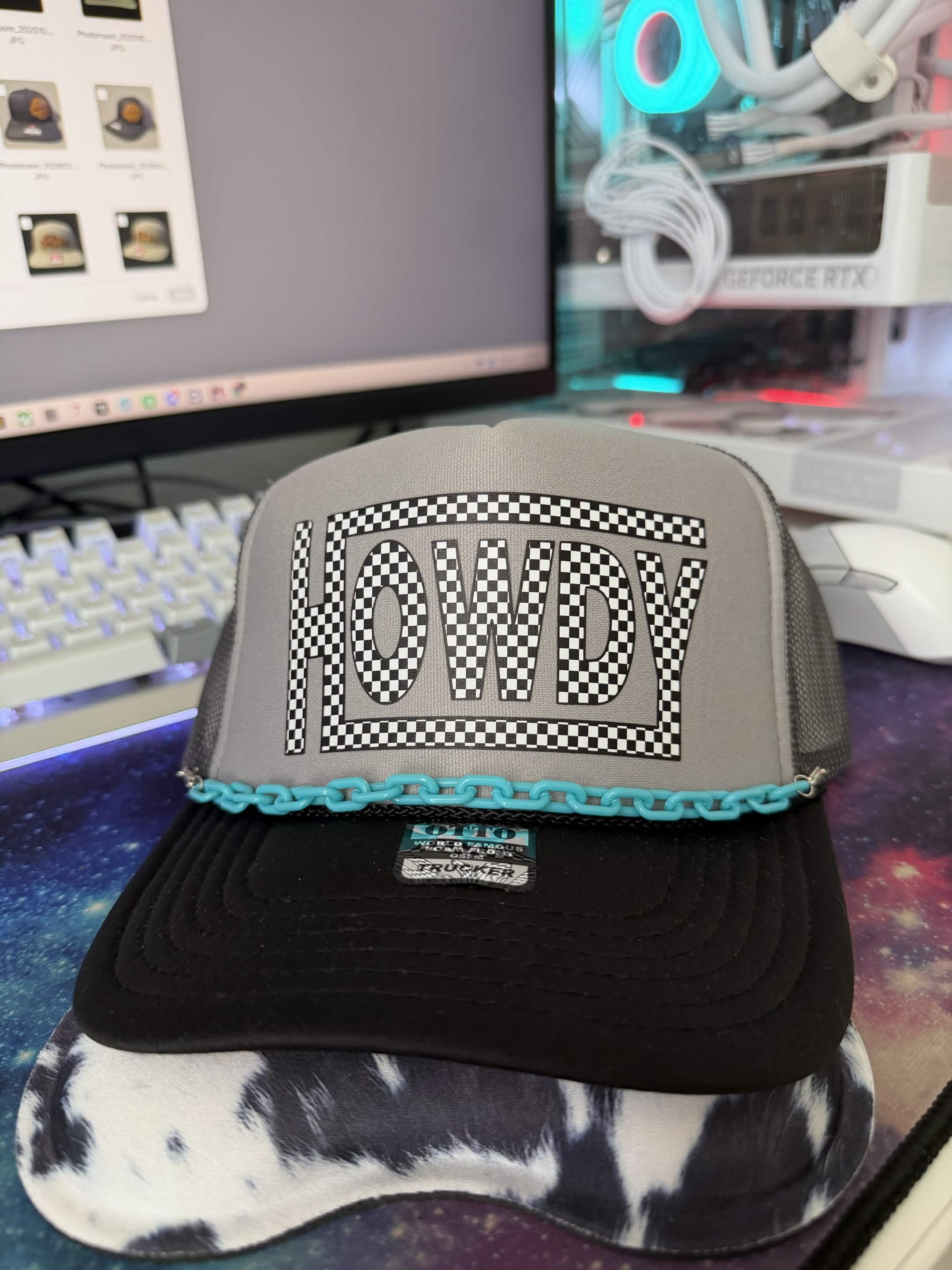 A gray and black trucker hat with a turquoise chain detail and a large checkered 'HOWDY' graphic on the front, displayed on a desk in front of a computer monitor.